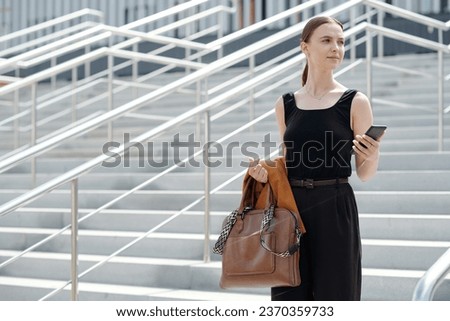 Young stylish woman in quiet luxury attire holding smartphone, folded blazer and brown leather handbag while standing against staircase