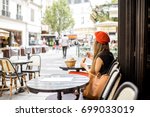 Young stylish woman in red beret having a french breakfast with coffee and croissant sitting oudoors at the cafe terrace