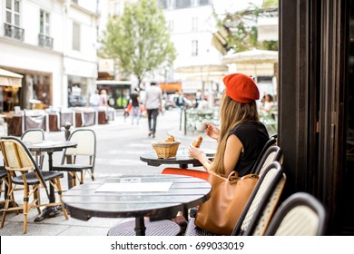 Young stylish woman in red beret having a french breakfast with coffee and croissant sitting oudoors at the cafe terrace - Shutterstock ID 699033019