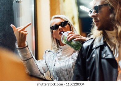 young stylish woman in sunglasses drinking mojito from plastic cup and showing middle finger at camera while spending time with boyfriend on street at night
