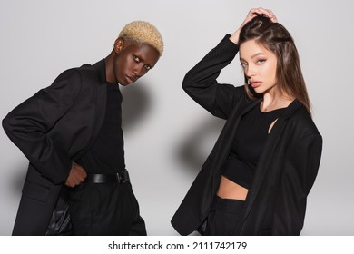 Young And Stylish Woman Touching Hair Near Blonde African American Man Posing With Hand On Waist On Grey With Shadow