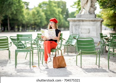 Young Stylish Woman Tourist In Red Cap And Pants Sitting With Paper Map On The Famous Green Chairs In Tuileries Garden In Paris