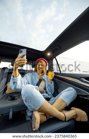 Young stylish woman uses smart phone while sitting relaxed with a coffee cup on backseat of car on the go. Wide angle view, modern car with panoramic rooftop