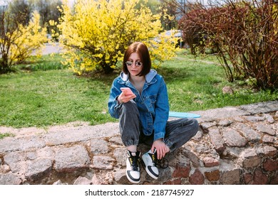 Young Stylish Woman Walks On Street With A Skateboard And Uses A Smartphone