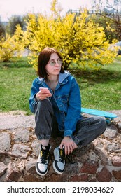 Young Stylish Woman Walks On Street With A Skateboard And Uses A Smartphone