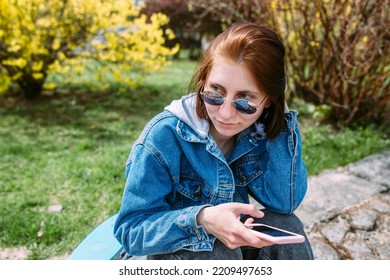 Young Stylish Woman Walks On Street With A Skateboard And Uses A Smartphone