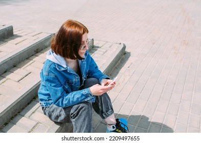 Young Stylish Woman Walks On Street With A Skateboard And Uses A Smartphone