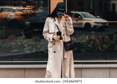 Young stylish woman wearing beige long seasonal trench coat walking around the city with a cup of coffee. Street style cloth concept