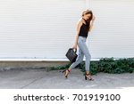 Young stylish woman wearing black cami top, blue cropped denim jeans, black high heel sandals and holding bag walking in the city street. Trendy casual outfit for summer or spring. Street fashion.