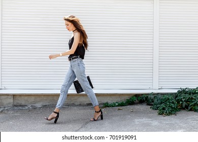 Young Stylish Woman Wearing Black Cami Top, Blue Cropped Denim Jeans, Black High Heel Sandals And Holding Bag Walking In The City Street. Trendy Casual Outfit For Summer Or Spring. Street Fashion.