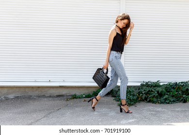 Young stylish woman wearing black cami top, blue cropped denim jeans, black high heel sandals and holding bag walking in the city street. Trendy casual outfit for summer or spring. Street fashion.