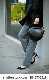 Young Stylish Woman Wearing Long Gray Jeans, Black High Heel Shoes And Black Silk Dress. She Is Holding Crocodile Textured Handbag. Street Style.