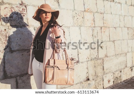 Young stylish woman wearing neutral blazer, hat and handbag walking on the city street in spring. Casual fashion, elegant look. Plus size model.