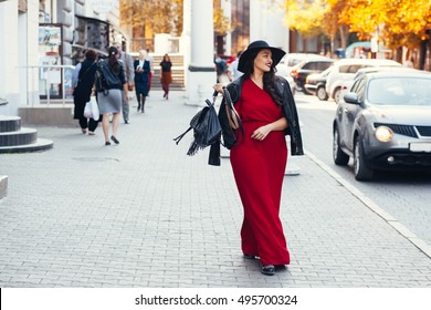 Young stylish woman wearing red maxi dress, black leather jacket and hat walking on the city street in autumn. Fall fashion, elegant look. Plus size model.