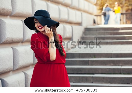 Young stylish woman wearing red maxi dress, black leather jacket and hat walking on the city street in autumn. Fall fashion, elegant look. Plus size model.