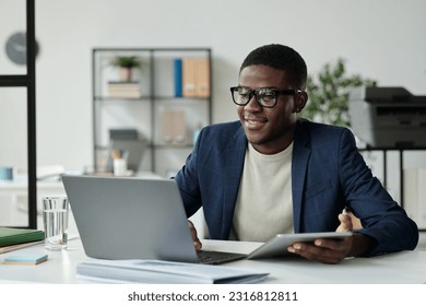 Young successful African American businessman in eyeglasses and elegant suit using mobile gadgets while sitting by workplace in front of laptop - Powered by Shutterstock