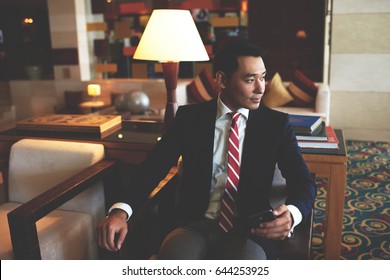 Young Successful Asian Businessman Dressed In Formal Wear Holding Mobile Phone While Sitting In Modern Office,confident Rich Men Entrepreneur Waiting For A Call On His Cell Telephone During Work Break