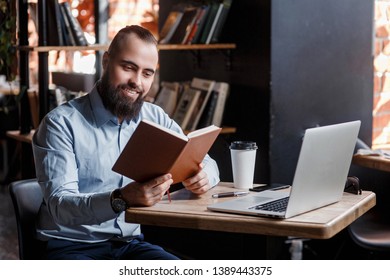 Young successful bearded businessman, working at a laptop, muzhschina looks at his notes in blaknote in a loft foise Coworking - Shutterstock ID 1389443375