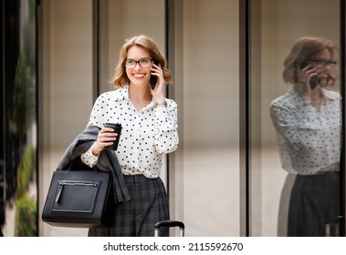 Young successful business lady in formal clothes with suitcase talking on mobile phone while standing on city street, smiling woman with coffee to go calling taxi by smartphone during business trip