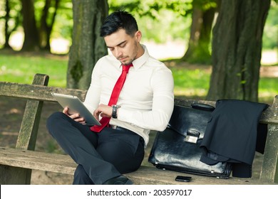 Young And Successful Business Man Sitting On The Park Bench Holding A Tablet And Checking Time