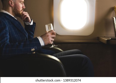 A young successful businessman in an expensive suit sits in the chair of a private jet with a glass of champagne in his hand and looks out the window