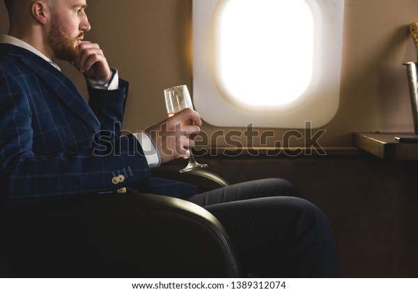 A young successful businessman in an
expensive suit sits in the chair of a private jet with a glass of
champagne in his hand and looks out the
window