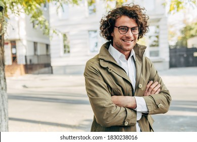 A Young Successful Businessman In Eyewear Smiling Broadly Posing Outdoors. Male Entrepreneur Resting In The City Street. Smart Guy In Casual Wears Spectacles With Curly Hair Walking Outside After Work