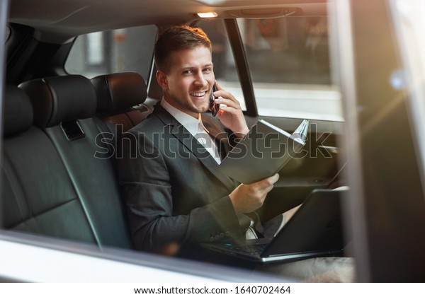 young successful\
businessman having phone conversation holds his planner while\
sitting in his car on the way to meetting with partners, looks\
happy, multitasking\
concept