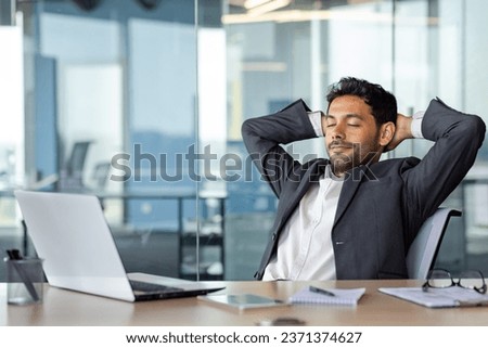 Young successful businessman resting dreaming and thinking inside office at workplace, man with hands behind head with closed glasses in business suit working with laptop.
