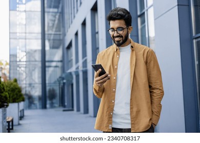 Young successful businessman walking in the city outside office building, hispanic smiling happy with achievement results, holding phone, using online application outdoors. - Powered by Shutterstock