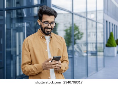 Young successful businessman walking in the city outside office building, hispanic smiling happy with achievement results, holding phone, using online application outdoors.