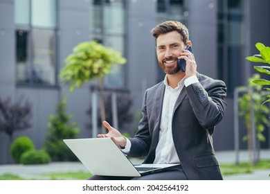 Young Successful Businessman Working On Laptop, Bearded Man In Business Suit Looking At Camera And Smiling, Talking On The Phone Near A Modern Office Center Outside