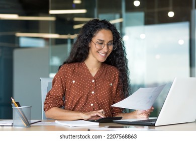 Young successful businesswoman behind paperwork, Hispanic woman working with documents and contracts inside modern office building, female worker using laptop, wearing glasses and curly hair smiling - Powered by Shutterstock