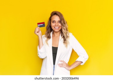 A young successful charming smiling blonde woman in a white jacket holding a plastic credit card in her hand isolated on a color yellow background. Shopping, payment for purchases, banking operations