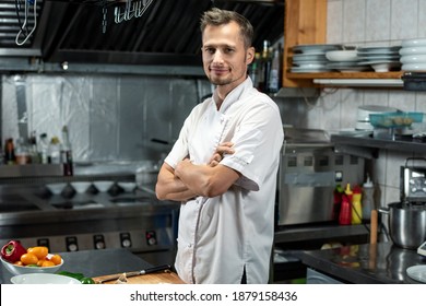 Joven y exitoso chef de un restaurante moderno con uniforme blanco que cruza los brazos en el pecho mientras se pone de pie en la mesa en la cocina antes de la ensalada de cocina
