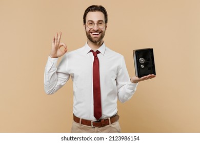 Young successful employee business man corporate lawyer 20s in white shirt red tie glasses work in office hold metal bank safe for money accumulation show ok gesture isolated on plain beige background
