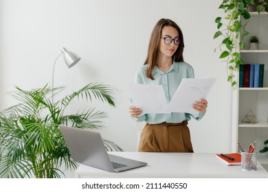 Young successful employee business woman in blue shirt hold paper account documents work stand at workplace white desk with laptop pc computer at light modern office indoors Achievement career concept - Shutterstock ID 2111440550