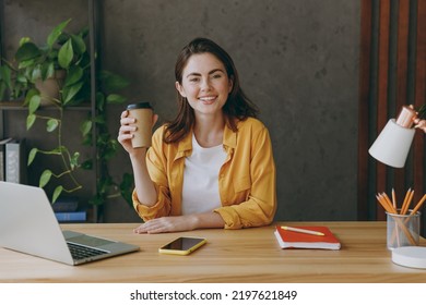 Young successful employee business woman wear casual yellow shirt hold takeaway delivery craft paper brown cup coffee to go sit work at wooden office desk with pc laptop. Achievement career concept - Powered by Shutterstock