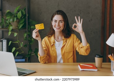Young successful employee business woman 20s wear casual yellow shirt hold in hand mock up of credit bank card show ok gesture sit work at wooden office desk with pc laptop. Achievement career concept - Shutterstock ID 2207973989