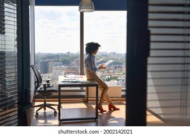 Young Successful Female African American Executive Manager Ceo Sitting On Desk Reading Financial Report In Modern Office In High Floor Corporate Building With Panoramic Urban Megapolis City View.