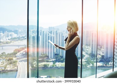 Young successful female boss is talking on cell telephone with customer and read information on website via digital tablet, while is standing in modern office interior near window with cityscape view