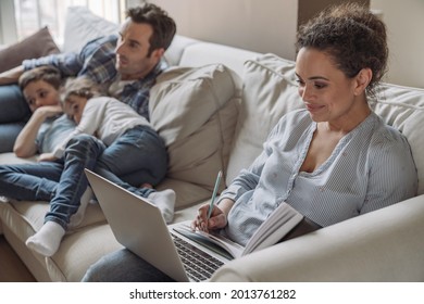 Young successful freelancer woman working at home sitting on sofa against the background of her husband and young son and daughter who are watching TV and having fun