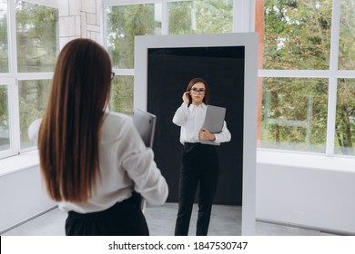 Young successful girl looks in the mirror. Girl holding a laptop. The girl is dressed in a white shirt and black pants