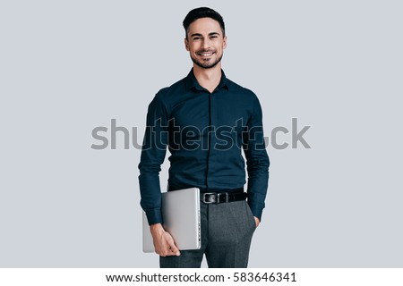 Young and successful. Good looking young man in shirt carrying laptop and looking at camera with smile while standing against grey background