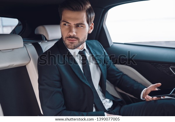 Young and successful. Handsome young\
man in full suit looking away while sitting in the car\
