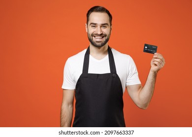 Young Successful Happy Fun Man 20s Barista Bartender Barman Employee Wearing Black Apron White T-shirt Work In Coffee Shop Hold Credit Bank Card Isolated On Orange Background. Small Business Startup.