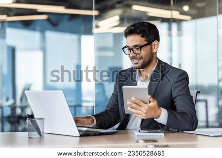 Young successful hispanic businessman at workplace using app on tablet computer, smiling man inside office building, working with laptop, wearing business suit, happy with achievement.