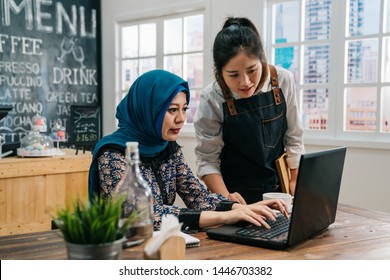 Young Successful Malay Woman Employee Small Business Owner Sitting In Morning Coffee Shop. Asian Girl Waitress Staff Talking With Friend Client Together Doing Project Giving Advice On Laptop Pc.