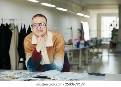 Young successful male fashion designer in smart casual attire and eyeglasses looking at camera while bending over workplace - Powered by Shutterstock