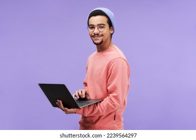 Young successful male freelancer or programmer in pink pullover, beanie hat and eyeglasses holding laptop and looking at camera - Shutterstock ID 2239564297